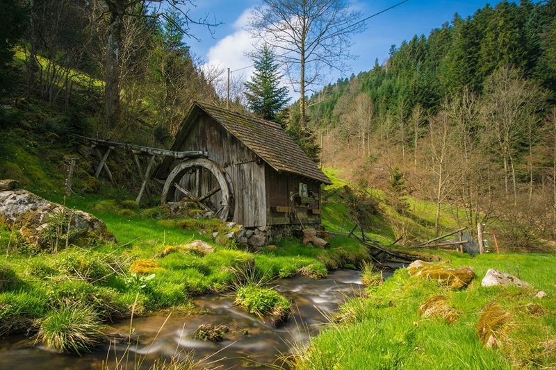 Historic wooden water mill with large wheel beside flowing stream in forested valley with moss-covered rocks