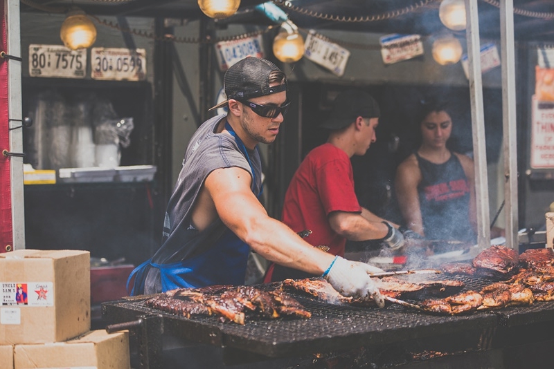 Street food vendor grilling meat at outdoor barbecue stand with smoke rising and license plates visible on wall
